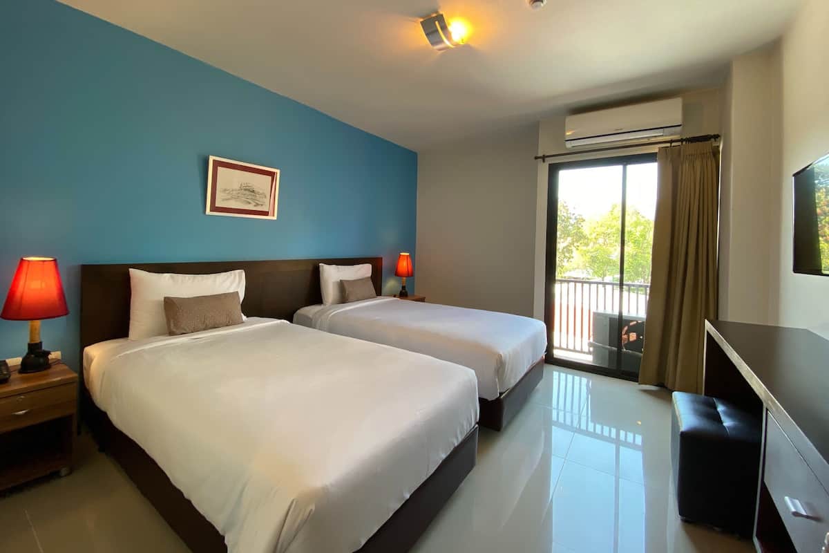 A hotel room with two beds, a desk, a TV, a window with curtains, and an air conditioning unit.