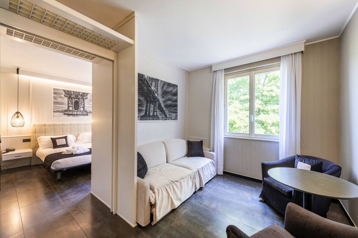 A modern hotel room with a bed, a sofa, a dining table, and a large window with curtains.