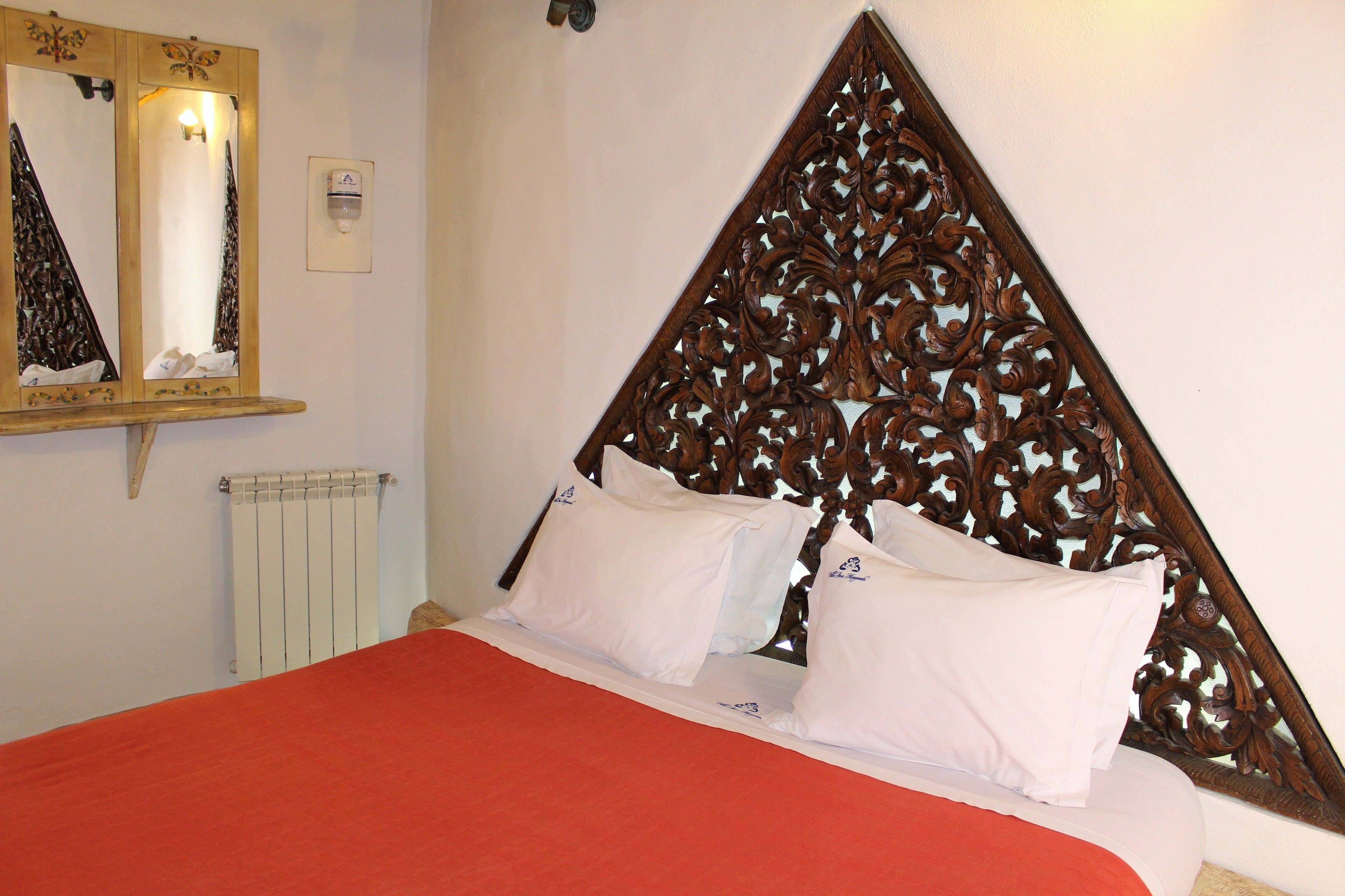 Ein Bett mit roter Bettdecke und weißen Kissen, ein hölzernes Kopfteil mit aufwendigen Schnitzereien, ein Spiegel über dem Bett und eine Wandleuchte.