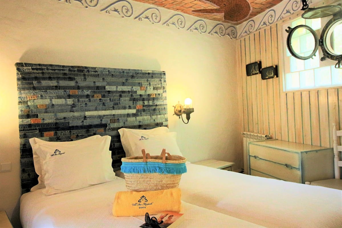 A hotel room with a bed featuring a denim headboard, a bedside table with a lamp, a small mirror, and a wooden dresser.