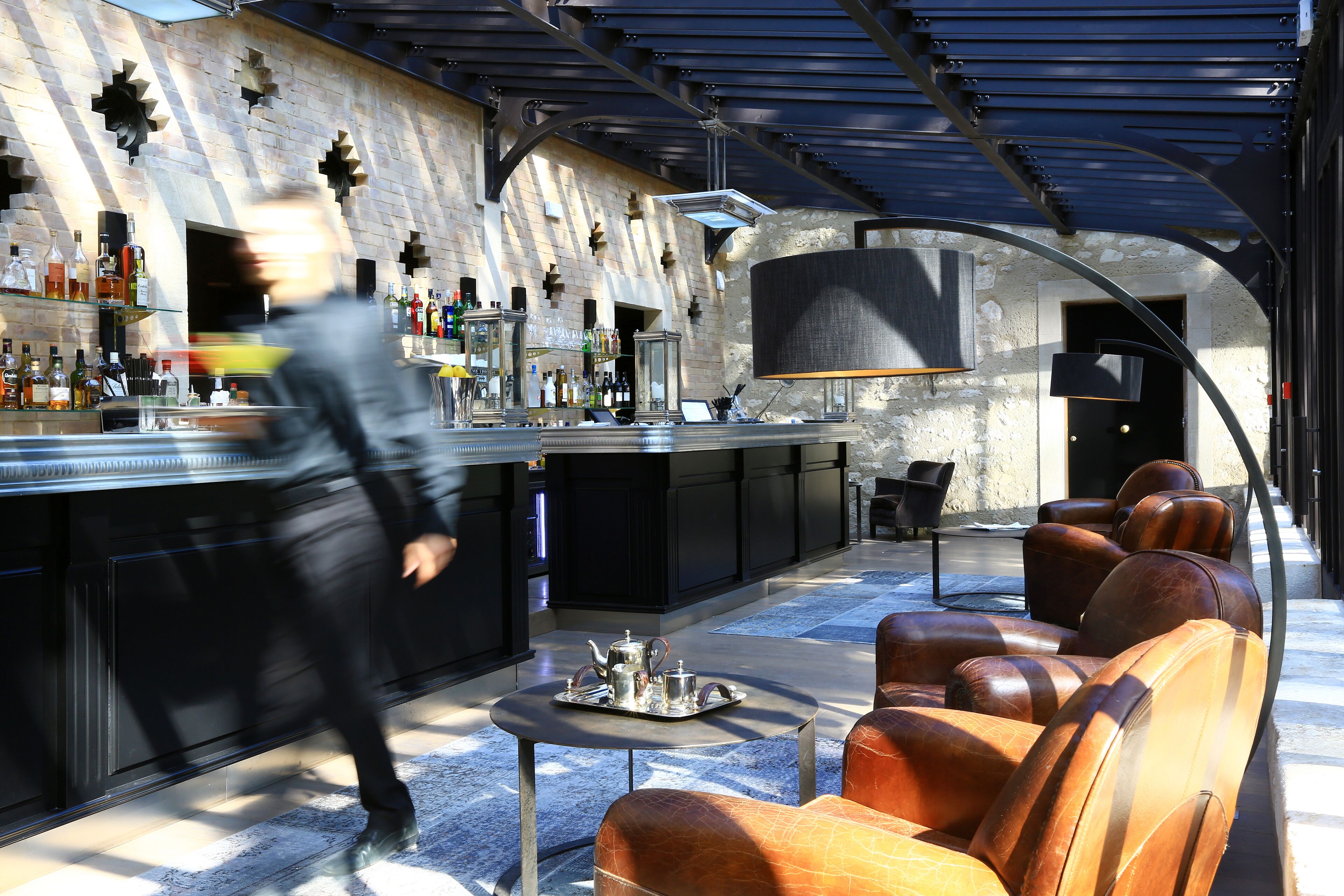 Zona de bar moderna con pared de piedra, barra con botellas y asientos de cuero.