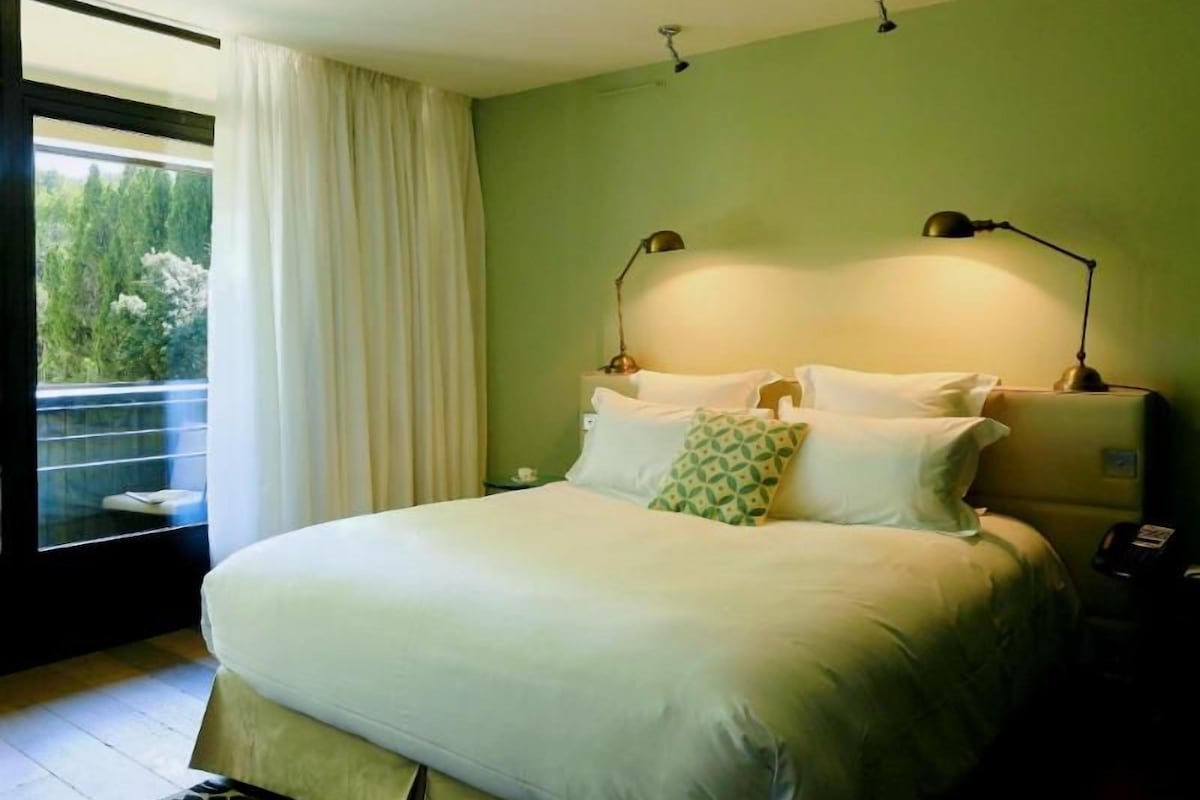 A bedroom with a large bed, a nightstand, a lamp, and a view of greenery outside.