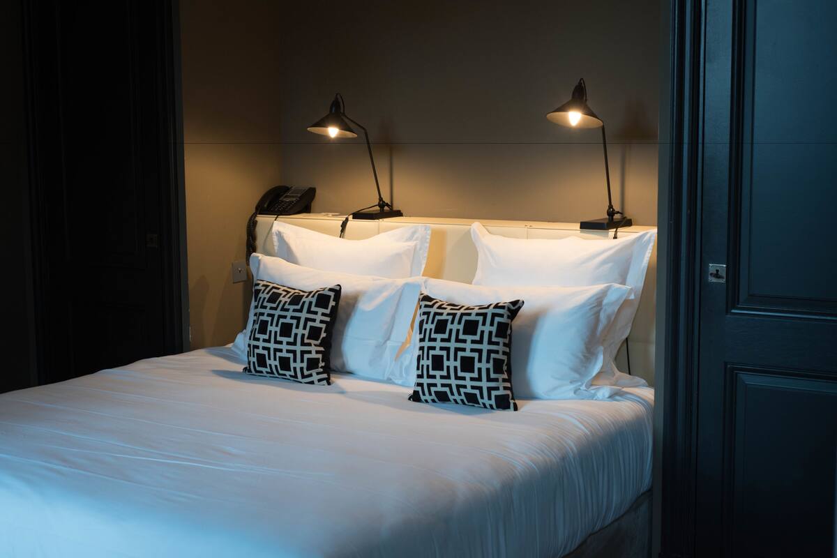 A neatly made bed with white linens and patterned pillows, two bedside lamps, and a telephone on the nightstand.