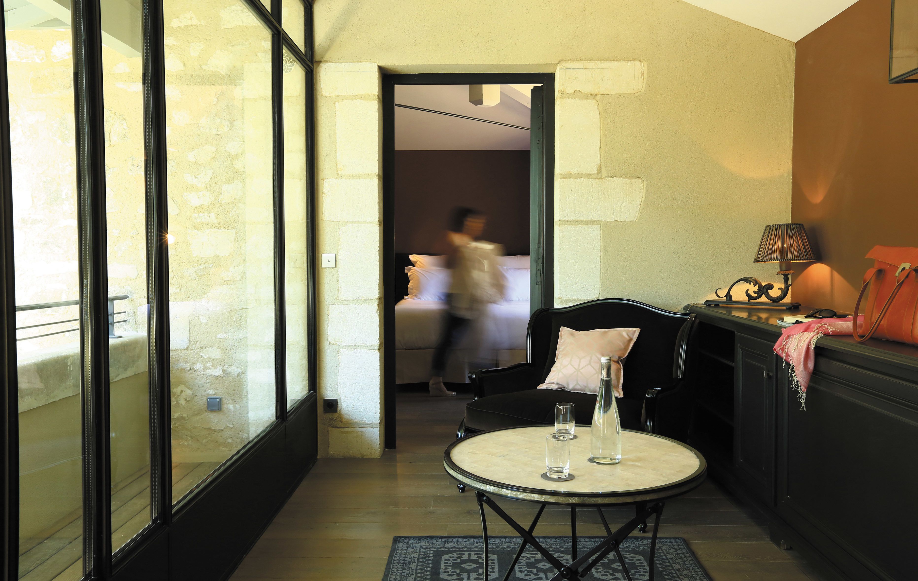 A hotel room with a glass partition, a round table with two glasses, a lamp, and a bed in the background.