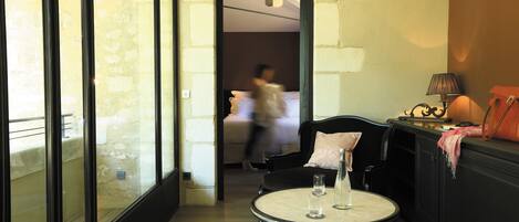 A hotel room with a glass partition, a round table with two glasses, a lamp, and a bed in the background.
