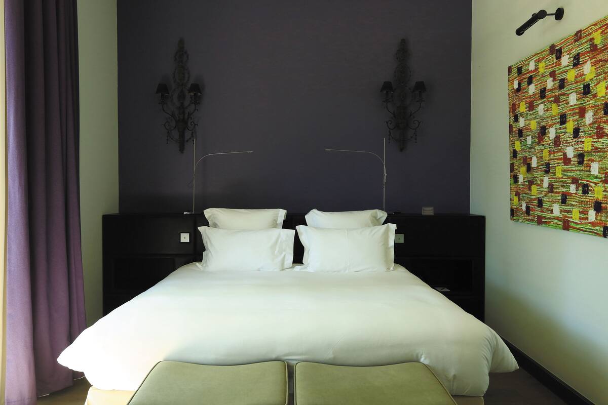 A neatly made bed with white linens, a dark headboard, and a wall-mounted lamp.