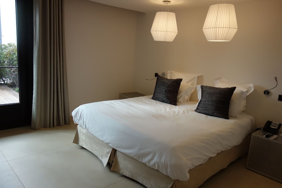 A hotel room with a large bed, two bedside lamps, a bedside table with a phone, and a view of the outdoors.