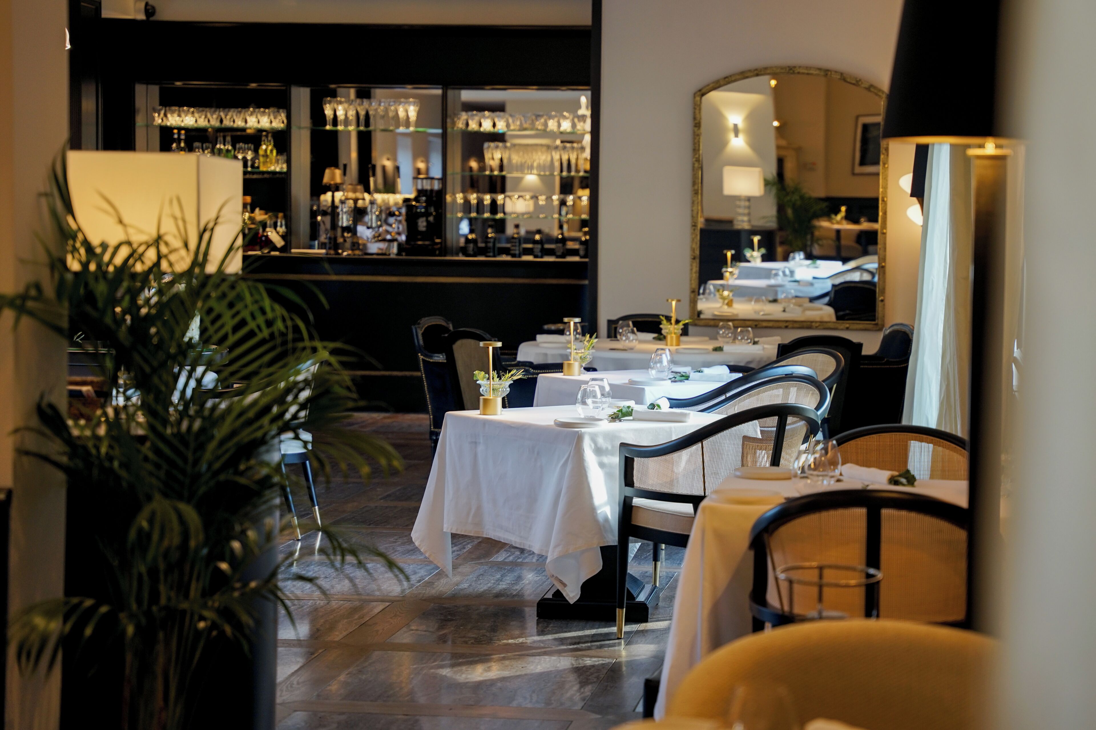 Ein Restaurant mit Barbereich im Hintergrund, Tische gedeckt mit weißen Tischdecken und Stühlen, und ein großer Spiegel an der Wand.