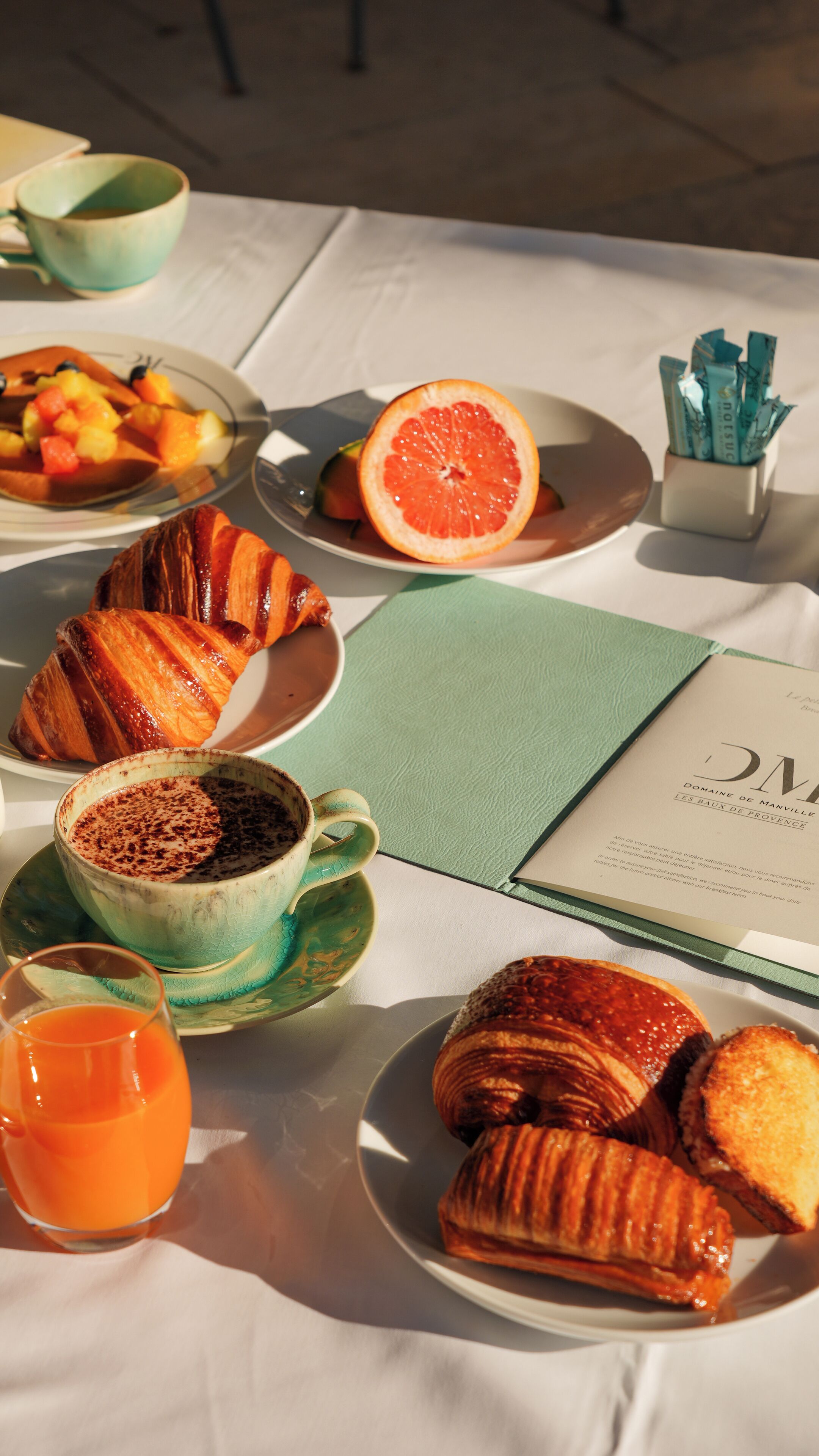 A table set with breakfast items including croissants, a grapefruit, a bowl of fruit, a cup of coffee, and a glass of orange juice.