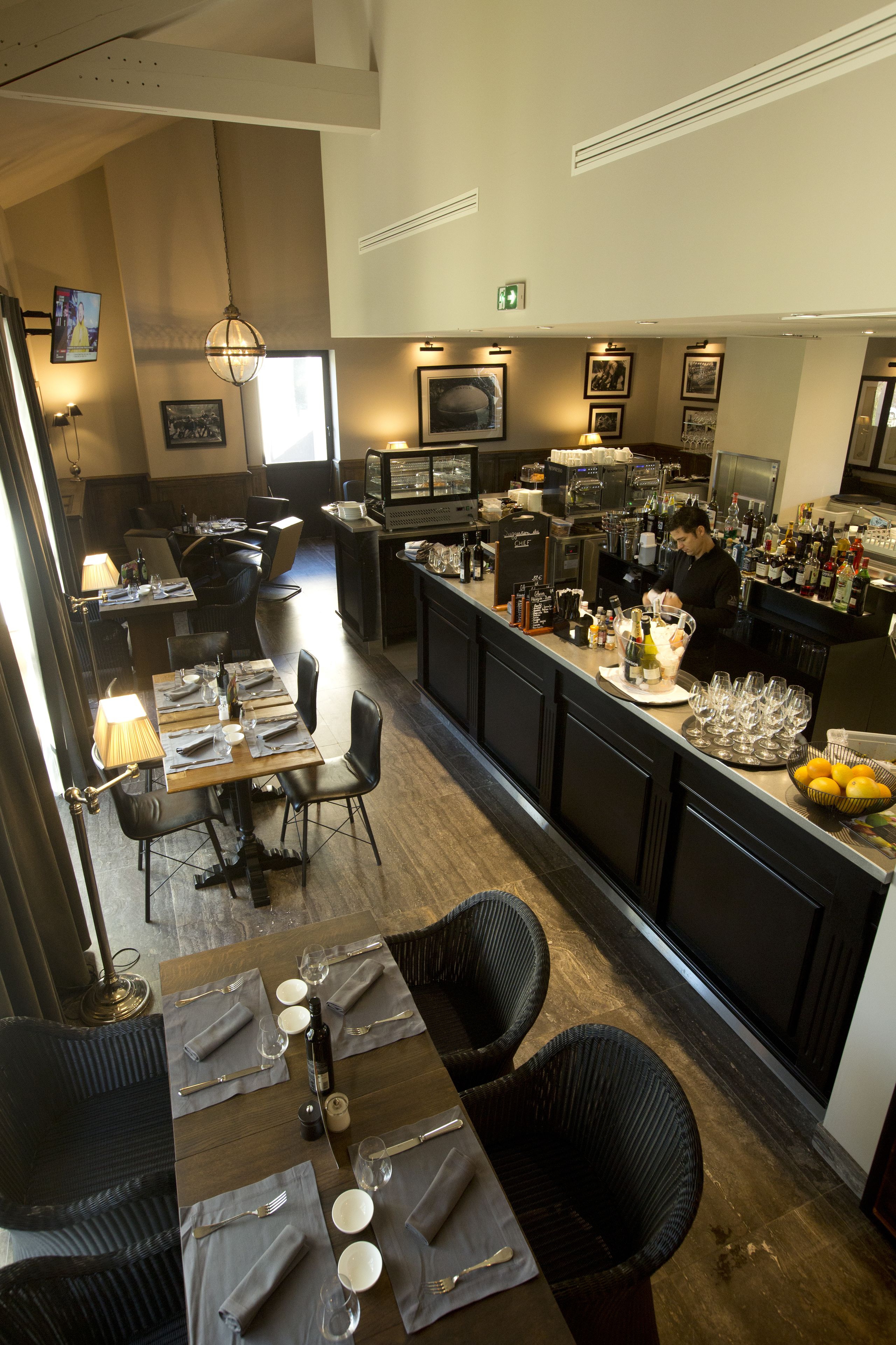 En restaurant med barområde, borde dækket med servietter, bestik og glas, samt en bar med en bartender der laver drinks.