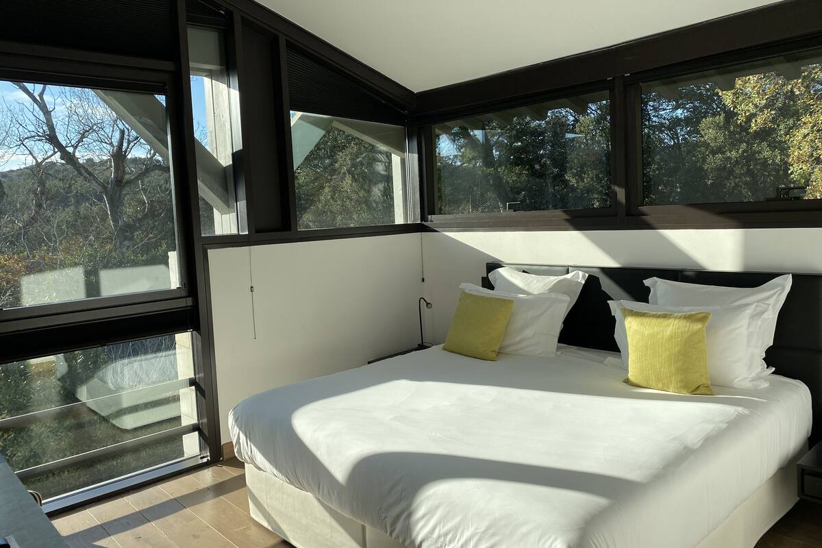 A modern bedroom with a large bed, multiple windows, and a view of trees.