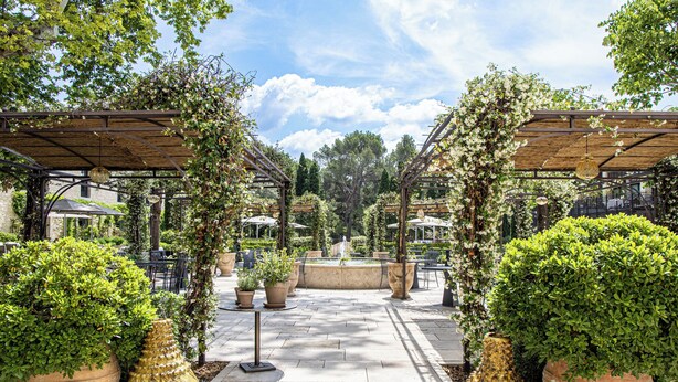 Un espace extérieur aménagé en terrasse, avec des tables et des chaises, entouré de plantes en pot et agrémenté d’une fontaine centrale.