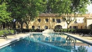 Une piscine avec des chaises longues, un bâtiment en pierre doté de nombreuses fenêtres et une fontaine au centre.