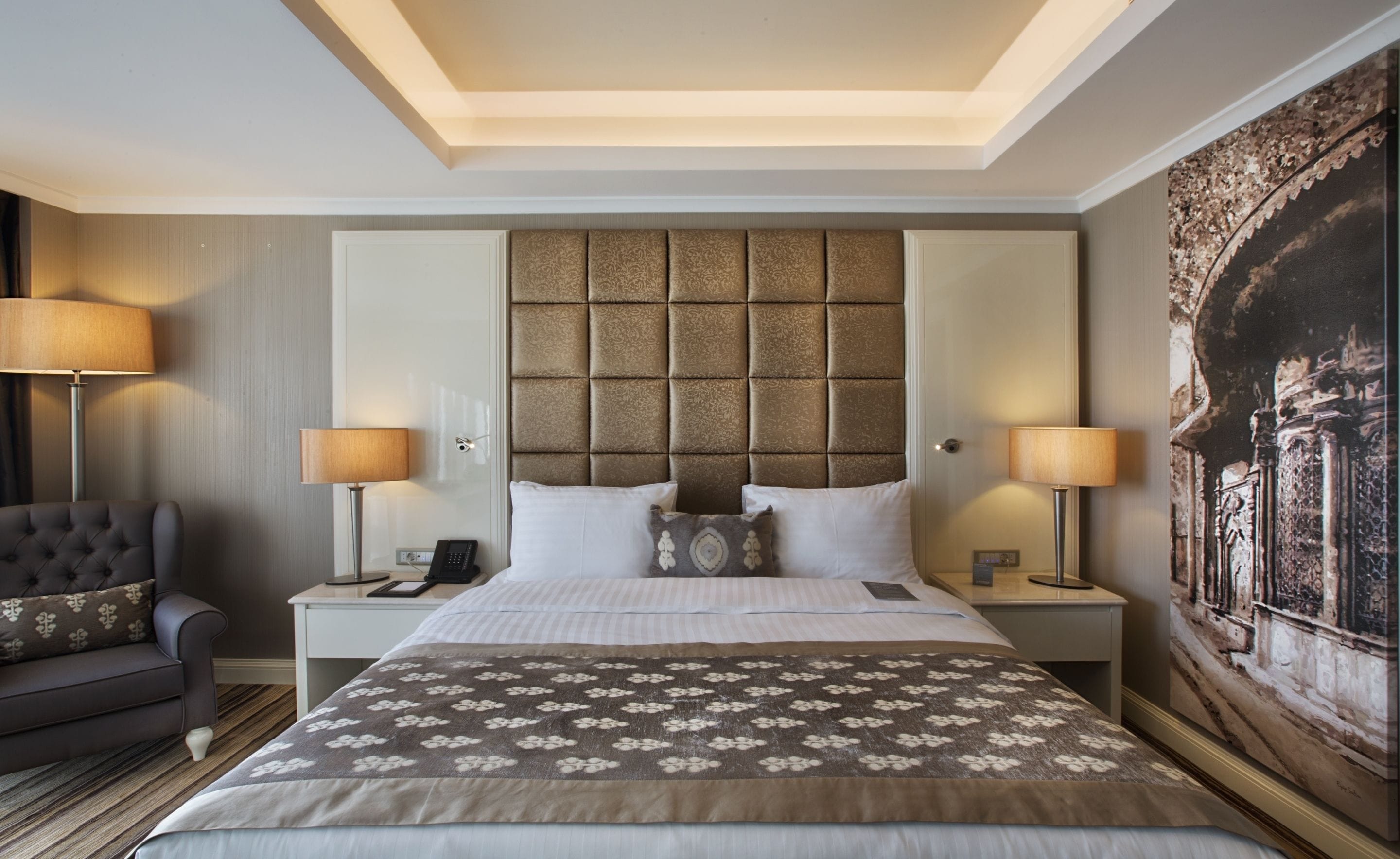 A hotel room with a large bed, a headboard with a textured panel, two bedside lamps, a nightstand with a phone, a sofa with a patterned throw, and a large painting on the wall.