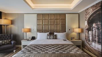 A hotel room with a large bed, a headboard with a textured panel, two bedside lamps, a nightstand with a phone, a sofa with a patterned throw, and a large painting on the wall.
