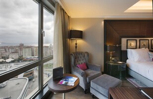 Una habitación de hotel con un ventanal grande, una cama, una silla, una mesita pequeña y una mesita de noche con lámpara.