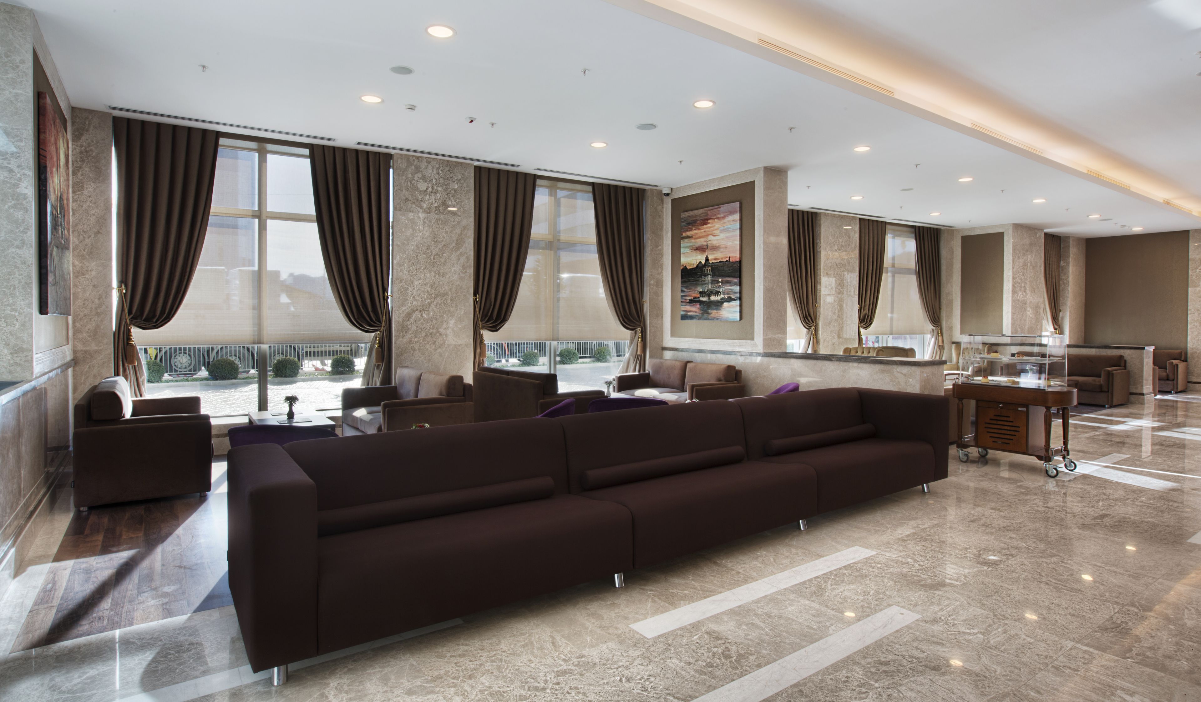 A modern hotel lobby with a large dark brown sofa, marble flooring, and large windows with curtains.