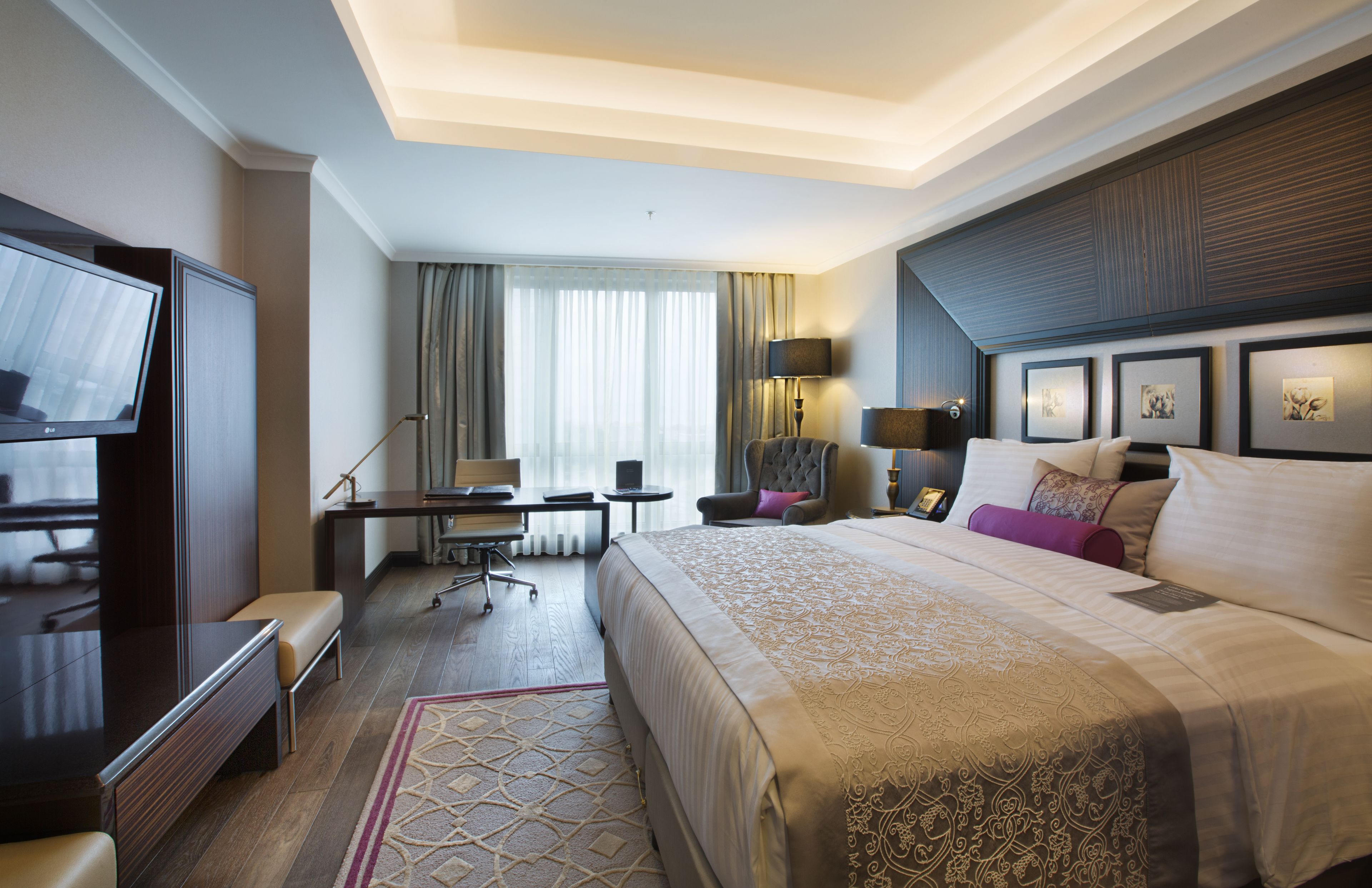 Ein Hotelzimmer mit einem großen Bett, einem Schreibtisch mit Stuhl, einem Fernseher und einem Fenster mit Vorhängen.