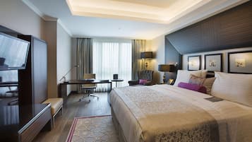 Ein Hotelzimmer mit einem großen Bett, einem Schreibtisch mit Stuhl, einem Fernseher und einem Fenster mit Vorhängen.