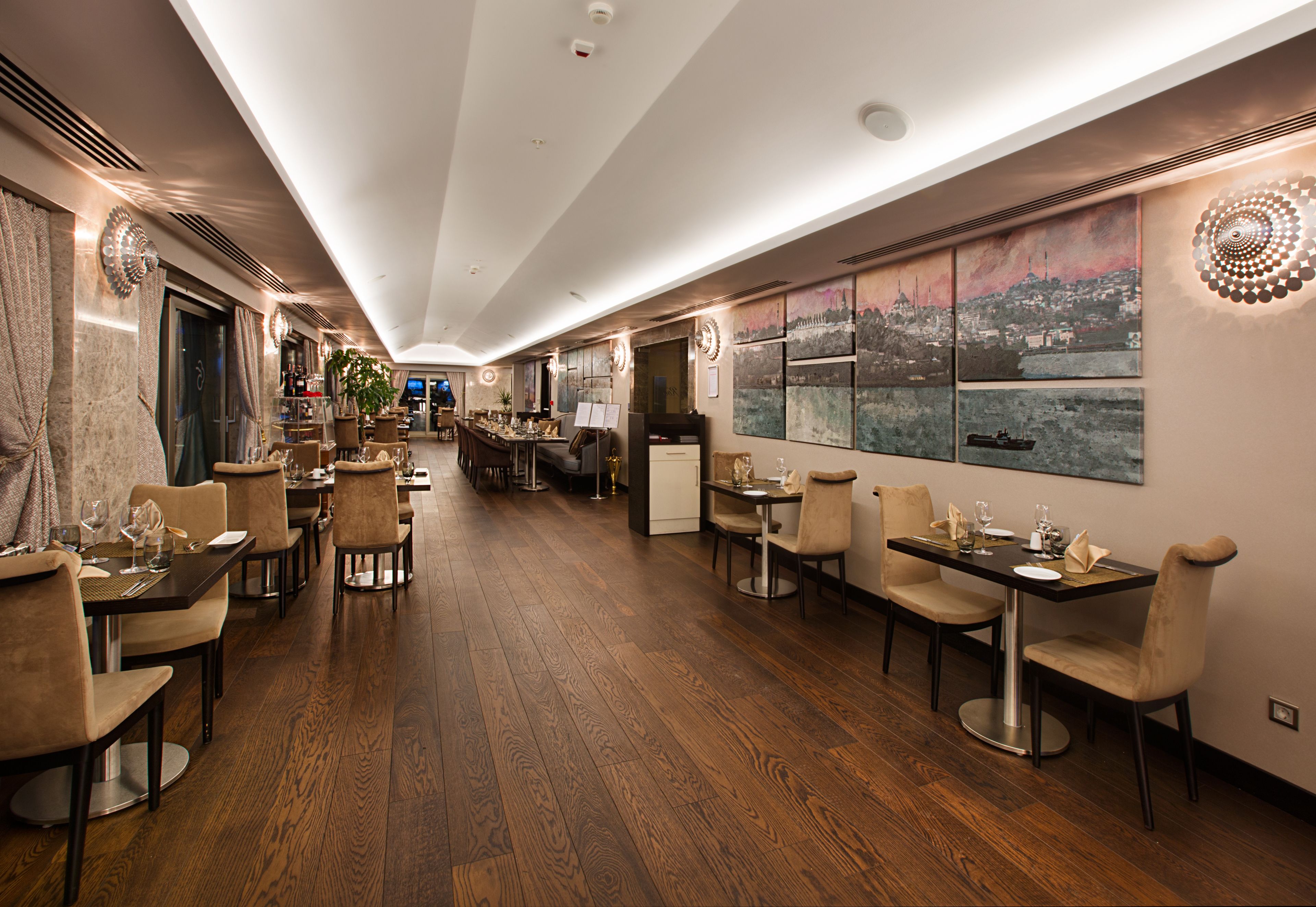 A restaurant with wooden flooring, multiple dining tables, and chairs. There are paintings on the walls and a large window with a cityscape view.