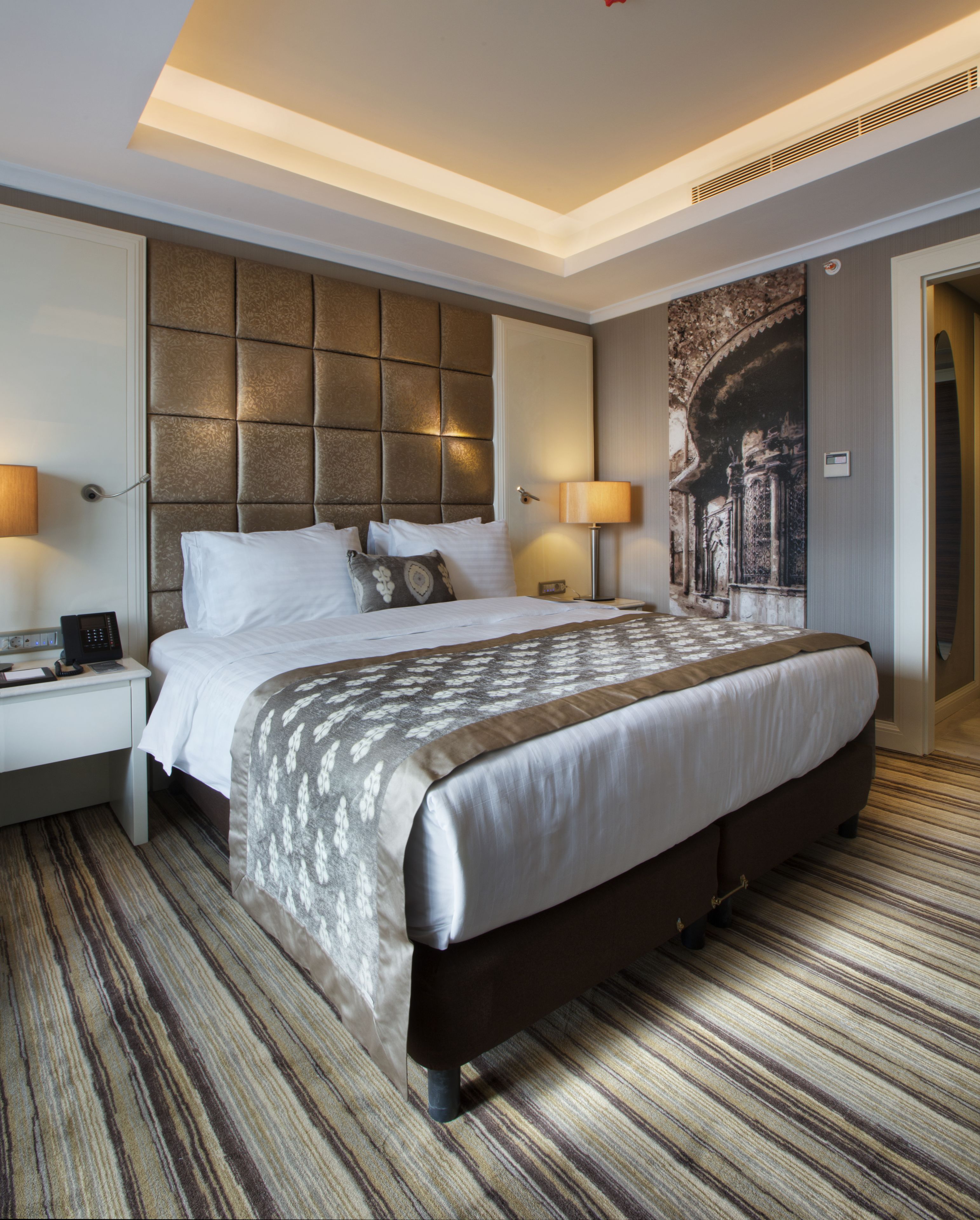 A hotel room with a large bed, a headboard, two bedside tables with lamps, and a carpeted floor.