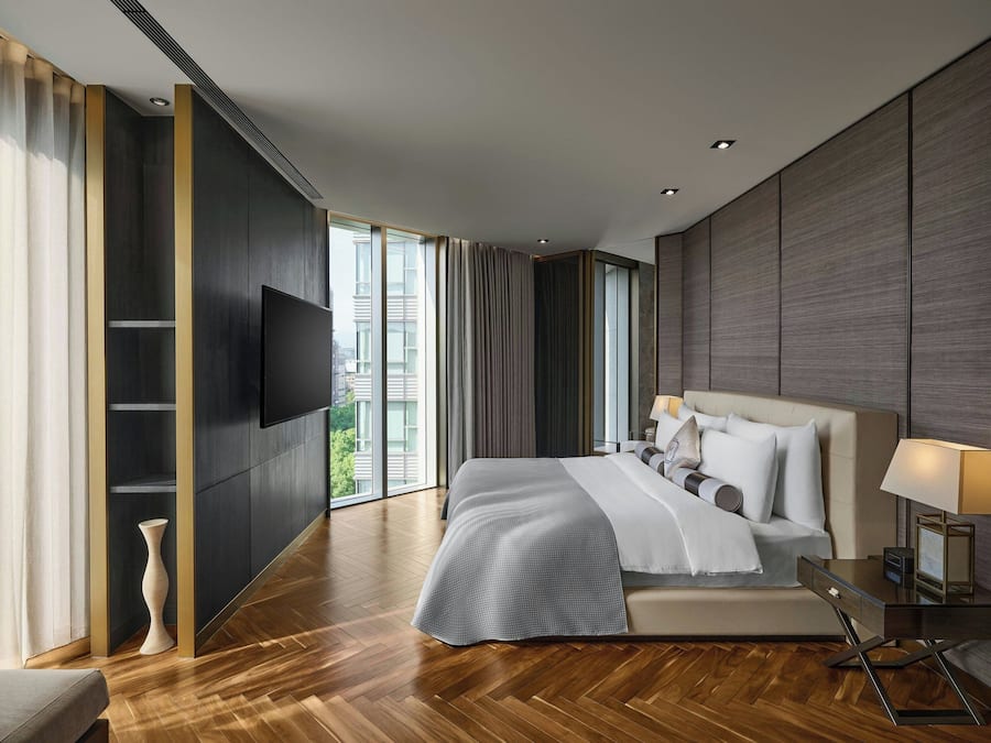 A modern hotel room with a large bed, a flat-screen TV, a bedside table with a lamp, and a view of the city through large windows.