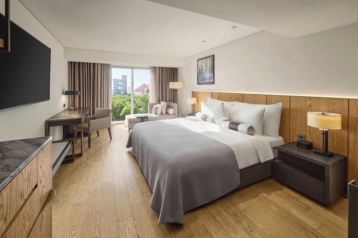 A hotel room with a large bed, a desk, a chair, a TV, and a view of the city.