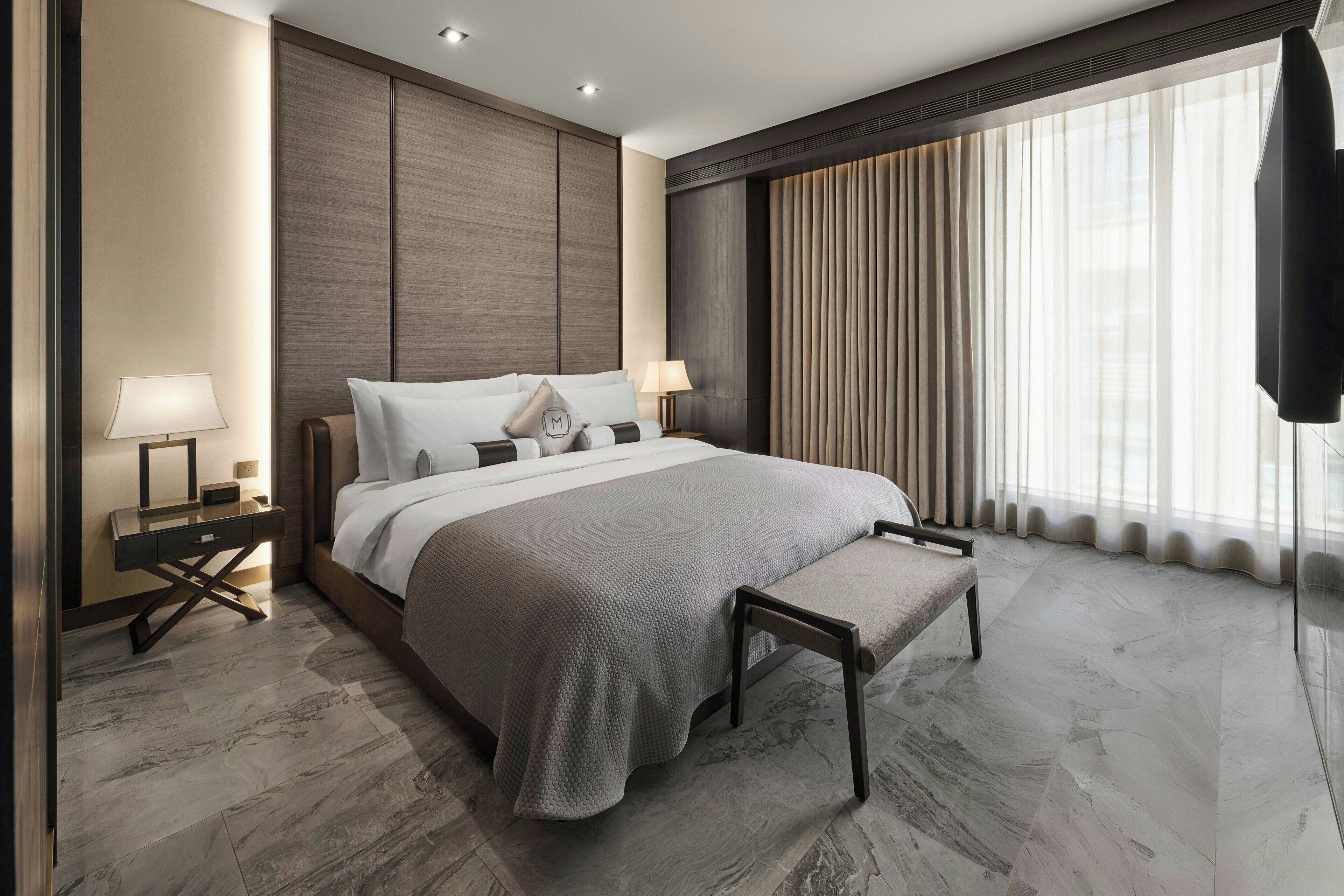 A modern hotel room with a large bed, two bedside tables with lamps, a desk, and a large window with curtains.