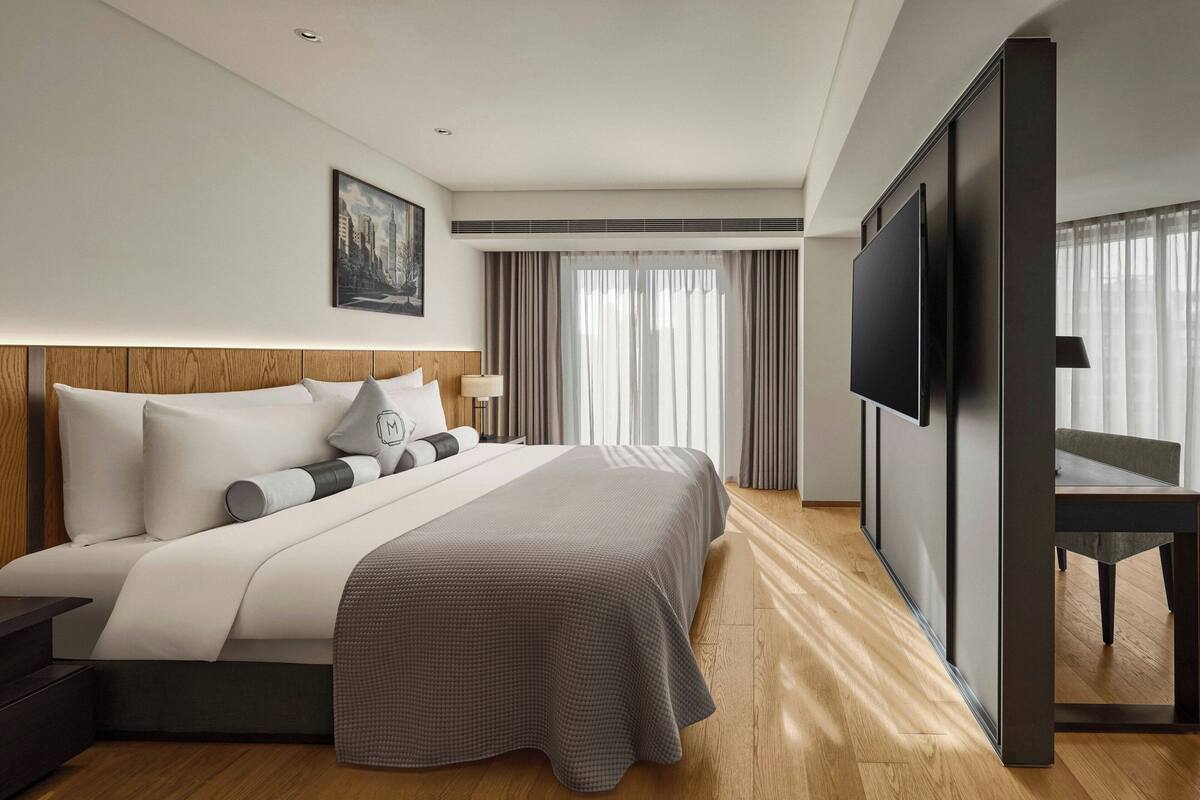 A modern hotel room with a large bed, a flat-screen TV, a wooden headboard, and a cityscape painting on the wall.