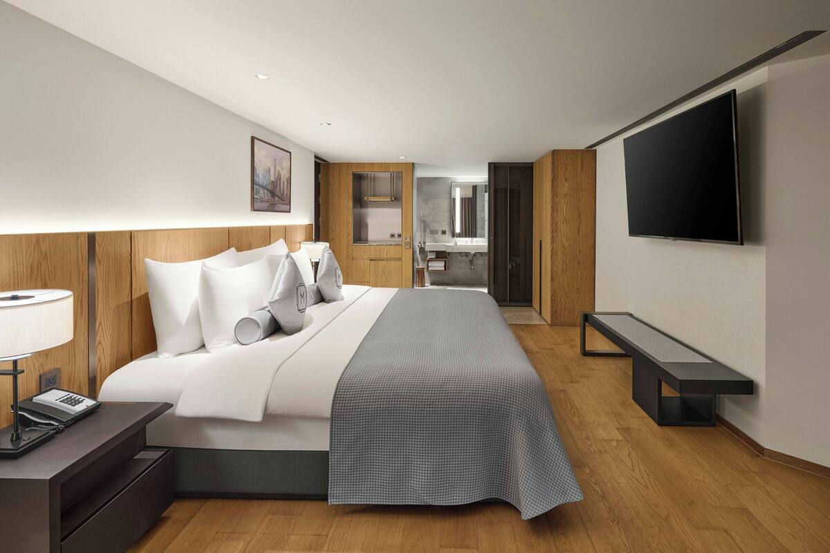 A modern hotel room with a large bed, bedside table with a lamp, a flat-screen TV mounted on the wall, and a wooden headboard.