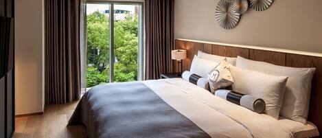 A hotel room with a large bed, a nightstand, a lamp, and a view of greenery through the window.