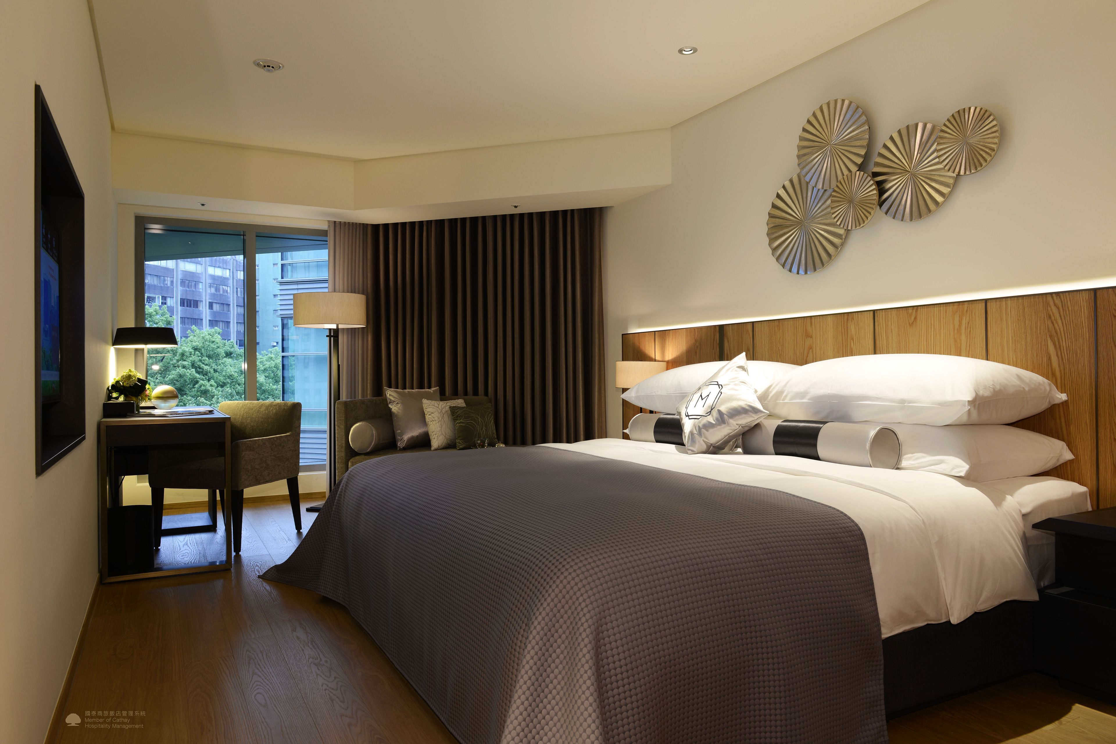 A modern hotel room with a large bed, a desk, a chair, and a view of the city through large windows.