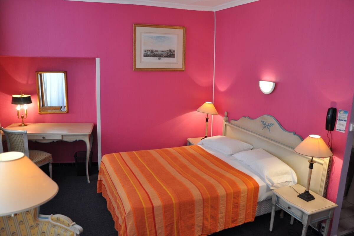 A hotel room with a bed, a desk, a chair, and a nightstand with lamps.