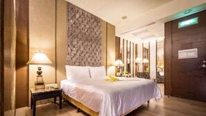 Una habitación de hotel con una cama grande, dos mesitas de noche con lámparas, un panel decorativo en la pared y una puerta de madera con letrero de salida de emergencia.
