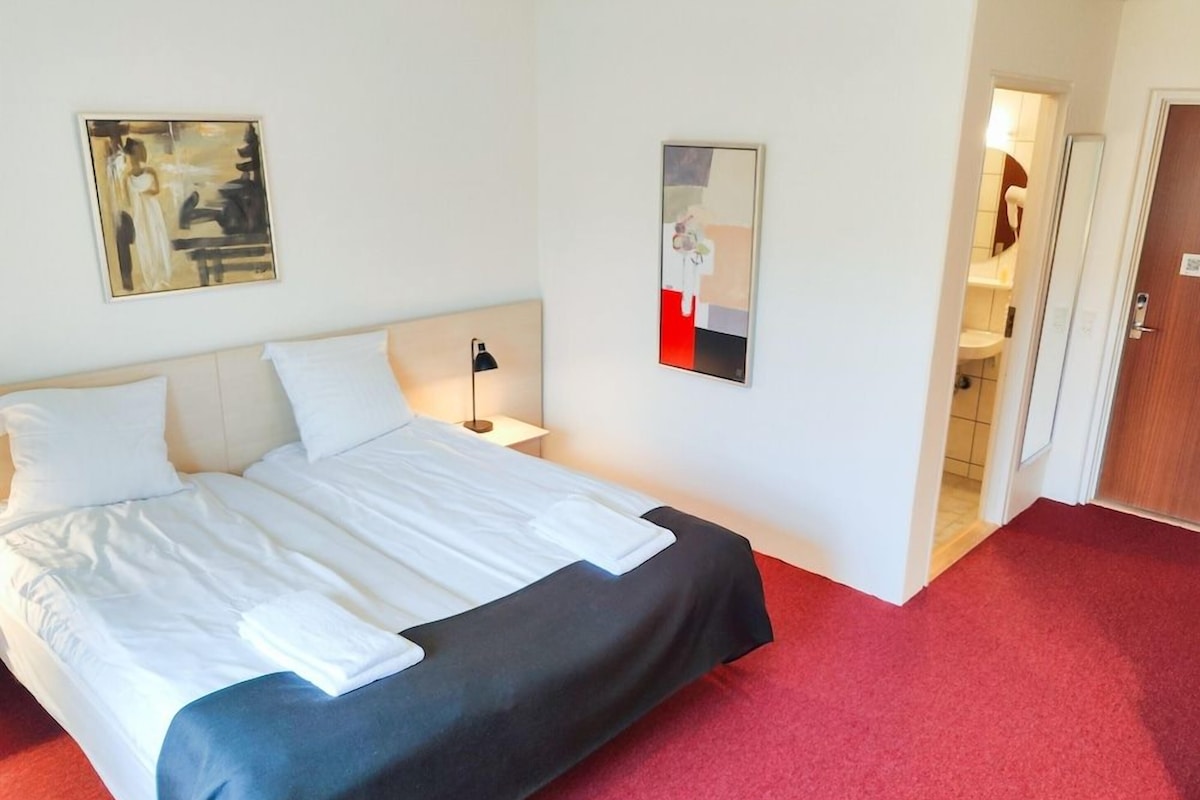 A hotel room with a bed, a nightstand, a lamp, and two paintings on the wall.