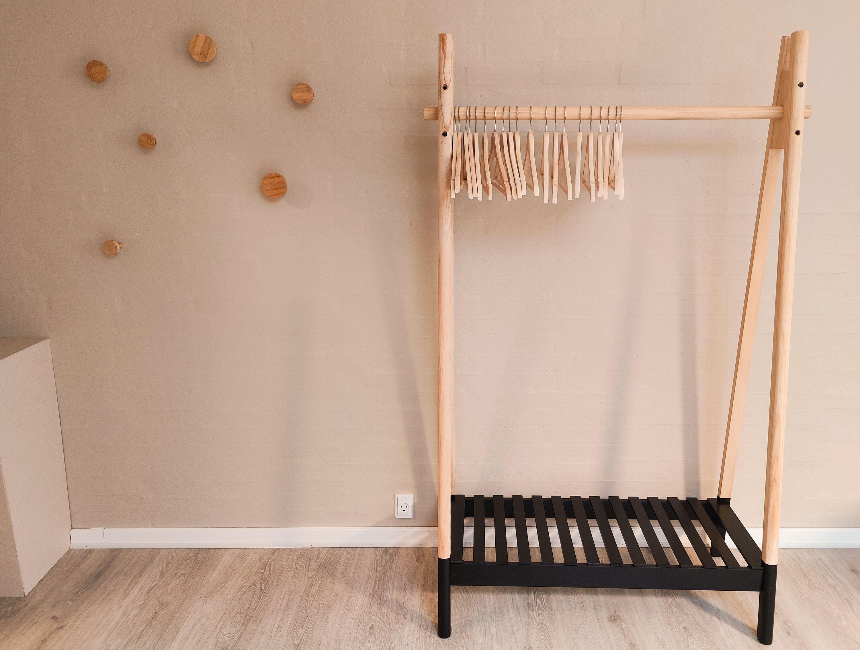 A wooden drying rack with pegs and a black shelf, against a beige wall with wooden decor.