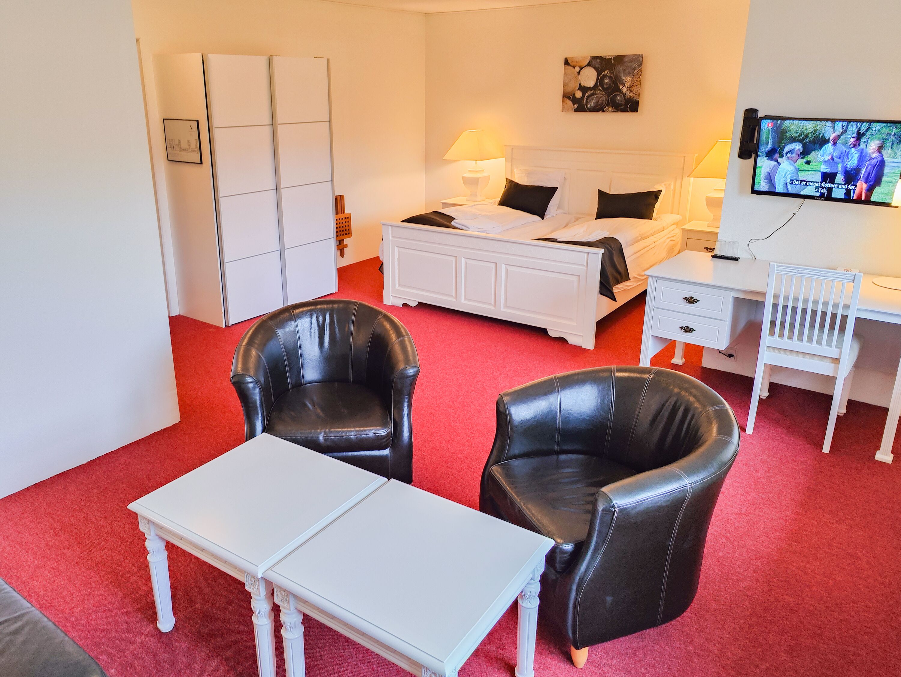 A hotel room with a bed, two black leather chairs, a white table, a desk, and a television on the wall.