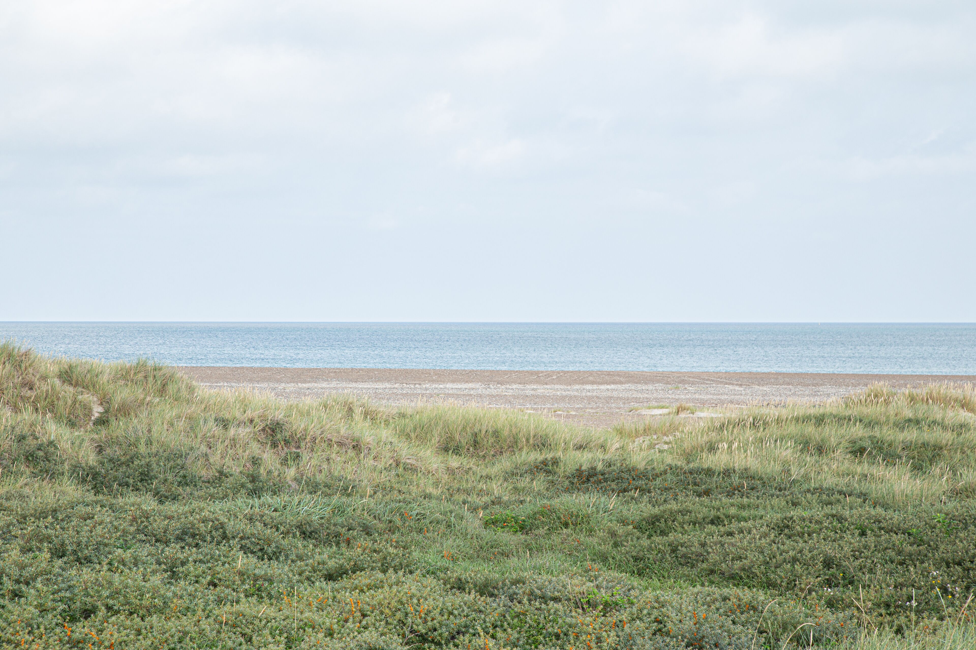 Une plage bordée de dunes herbeuses et une mer calme sous un ciel nuageux.