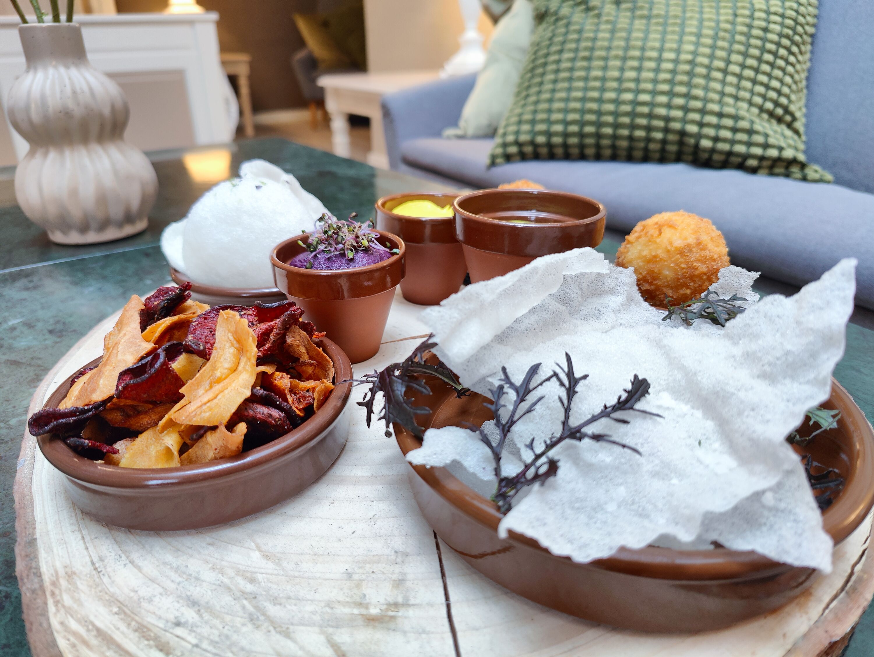 Una mesa con varios platos, incluyendo un cuenco de papas fritas, un cuenco que parece ser un postre, y dos pequeños macetas de terracota con una sustancia púrpura y una sustancia amarilla.