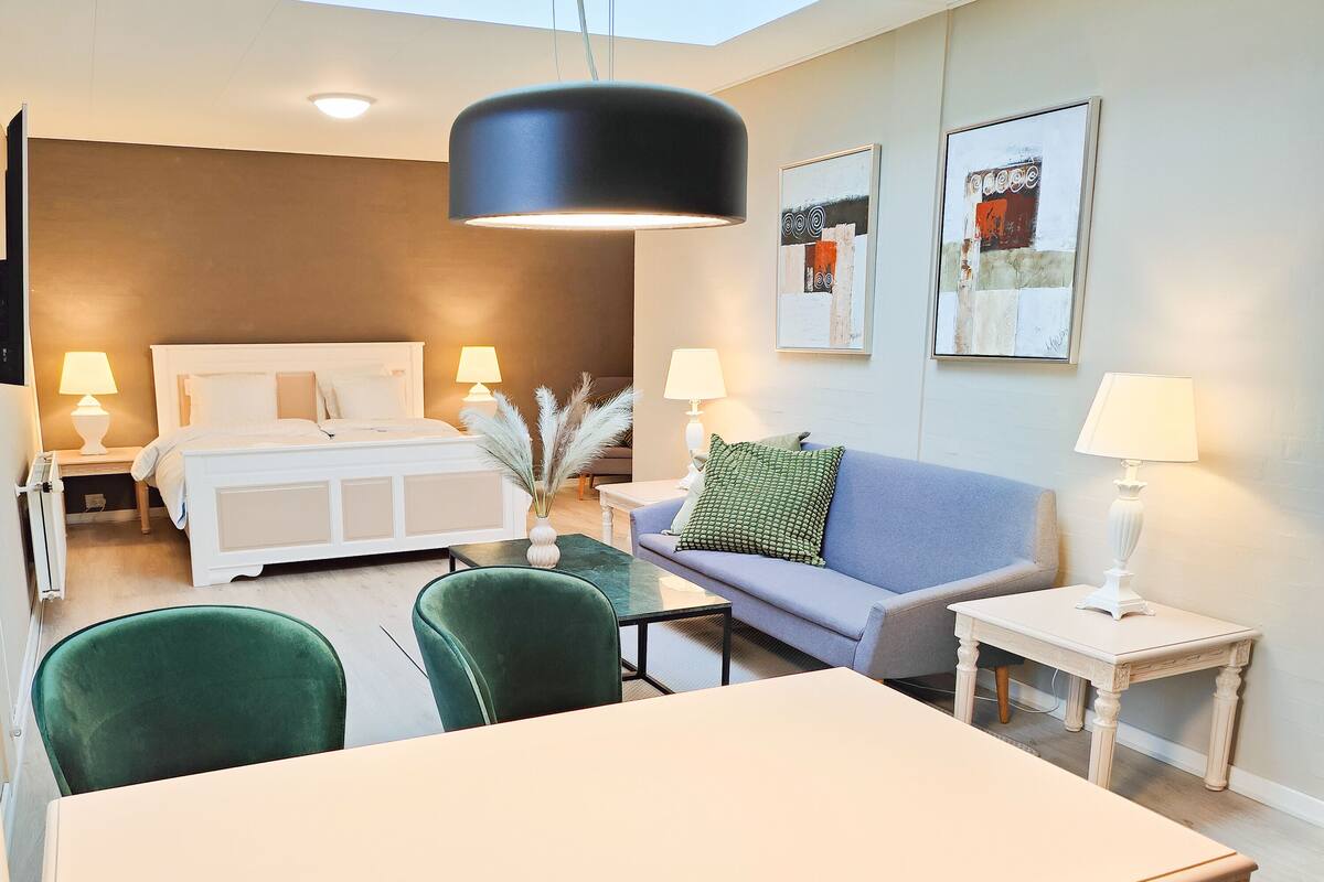 A hotel room with a bed, a sofa, a dining table, and two chairs. There are two bedside tables with lamps, a wall-mounted artwork, and a ceiling light.