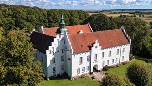 A large, white building with a red-tiled roof and a prominent tower, surrounded by lush greenery and fields. - Rønnes Hotel (Fjerritslev)