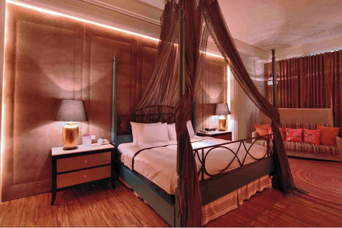A four-poster canopy bed with a wooden headboard, a nightstand with a lamp, and a sofa with cushions.