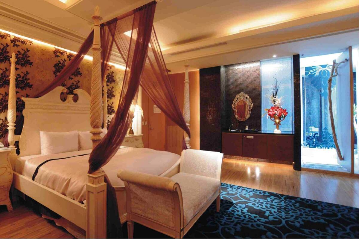 A four-poster bed with a canopy, a bedside table with a lamp, a chair, and a large mirror on the wall.