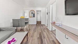 A modern living room with a grey sofa, a dining table with chairs, a TV mounted on the wall, and a hallway leading to a bathroom. - 5000 Motel (Penticton)