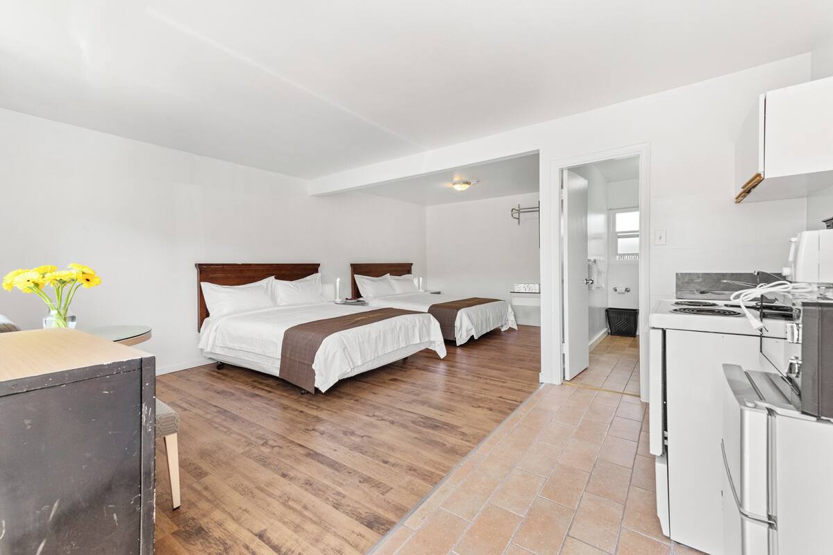 Una camera d'albergo con due letti, un angolo cottura e un bagno visibile attraverso una porta aperta.