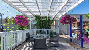 An outdoor patio with a glass table and chairs, surrounded by a white fence, and a playground in the background. - 5000 Motel (Penticton)