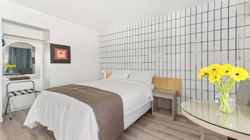 Habitación de hotel con una cama, una mesita de noche, una silla, una pequeña mesa con un florero y un papel tapiz con diseño de cuadrícula.