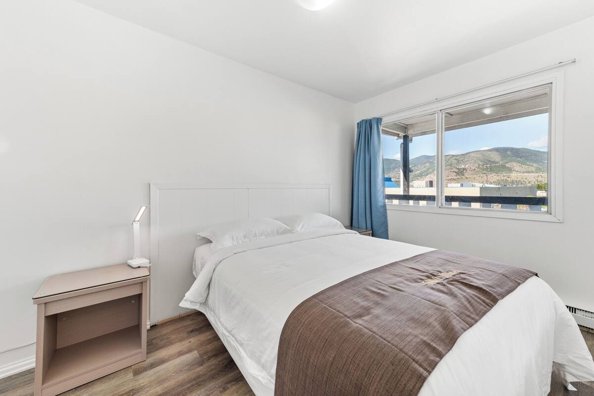 Una camera da letto con un letto, un comodino, una lampada, una finestra con vista sulle colline e una tenda.