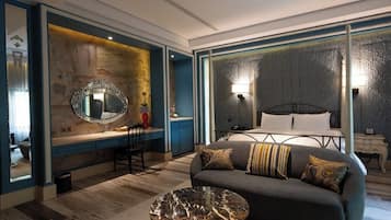 A modern hotel room with a sofa, a round coffee table, a large mirror, and a bed with a headboard.