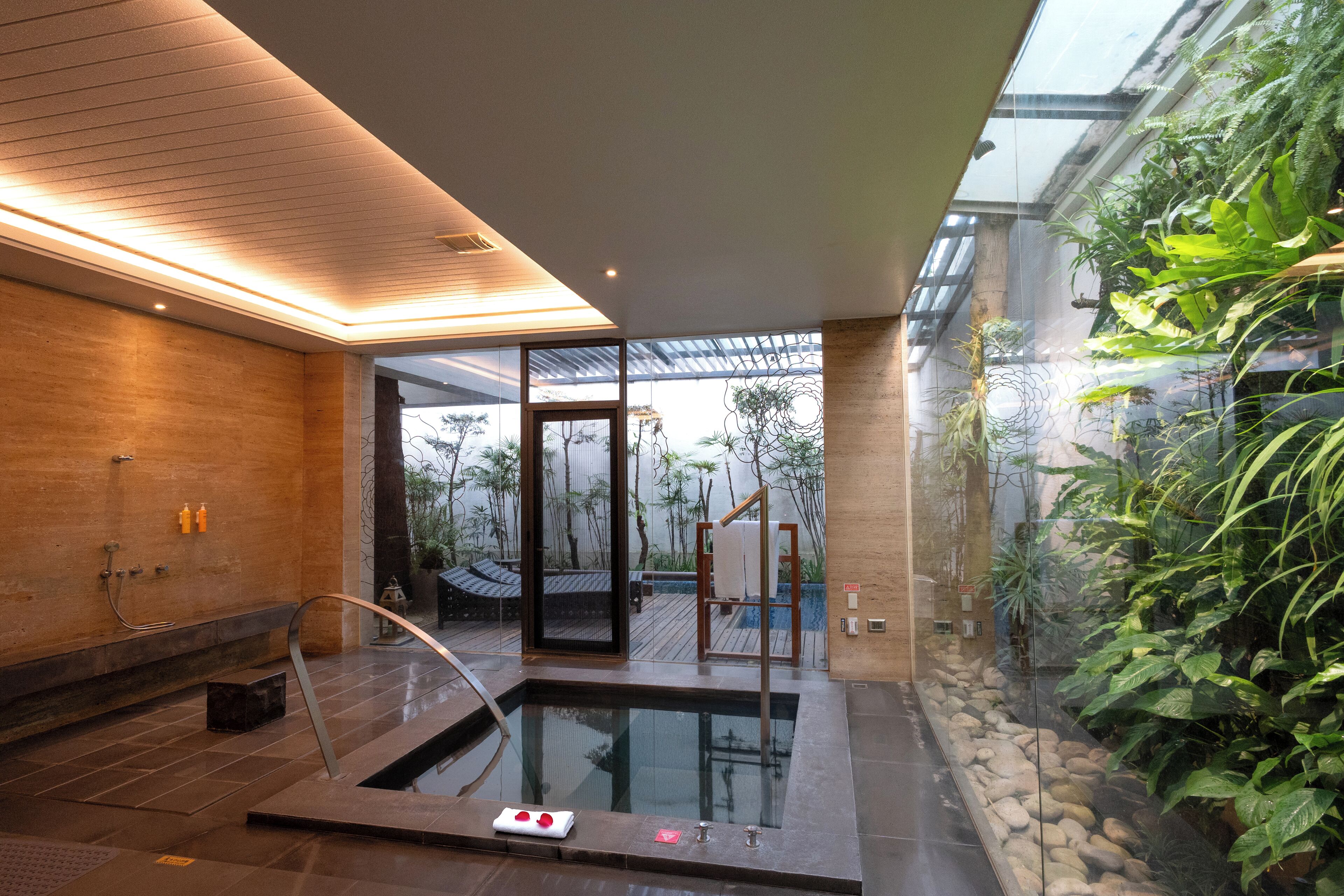 A modern spa area with a glass-enclosed hot tub, wooden flooring, and a view of a tropical garden.